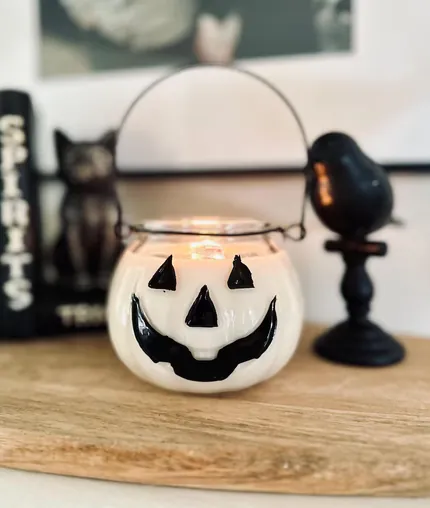Lit pumpkin lantern candle with a black jack-o-lantern face and wire handle